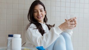 Smiling woman holding a glass while seated, with pill bottles on the table.